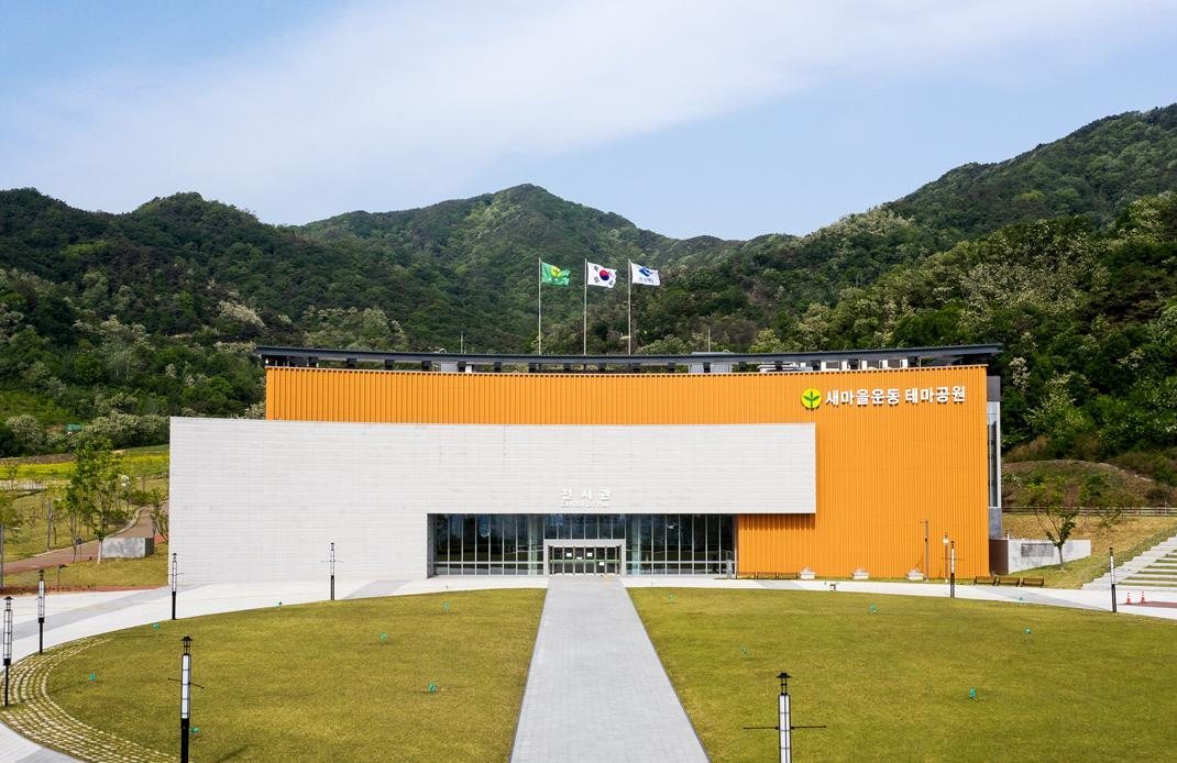 새마을운동테마공원 전시관 정면 — 주황색 외벽 건물과 앞 잔디 광장, 뒤로 산